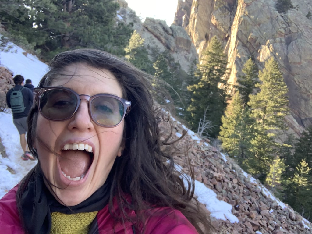 A person is taking a selfie with their mouth wide open in an expression of excitement or jubilation. The individual is wearing oversized sunglasses, a bright pink jacket, a yellow garment underneath, and a black neck accessory. Their hair is swept by the wind. In the background, there is a snow-covered trail on a hilly terrain with coniferous trees and rocky outcrops. There are other hikers walking on the trail in the middle distance, one wearing a gray backpack.
